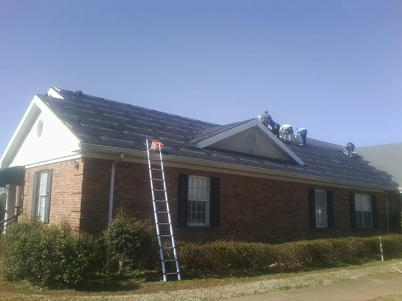 Crew tearing shingles off a brick ranch roof with a ladder set — Emergency Roof Repair work by our crew in Hansen