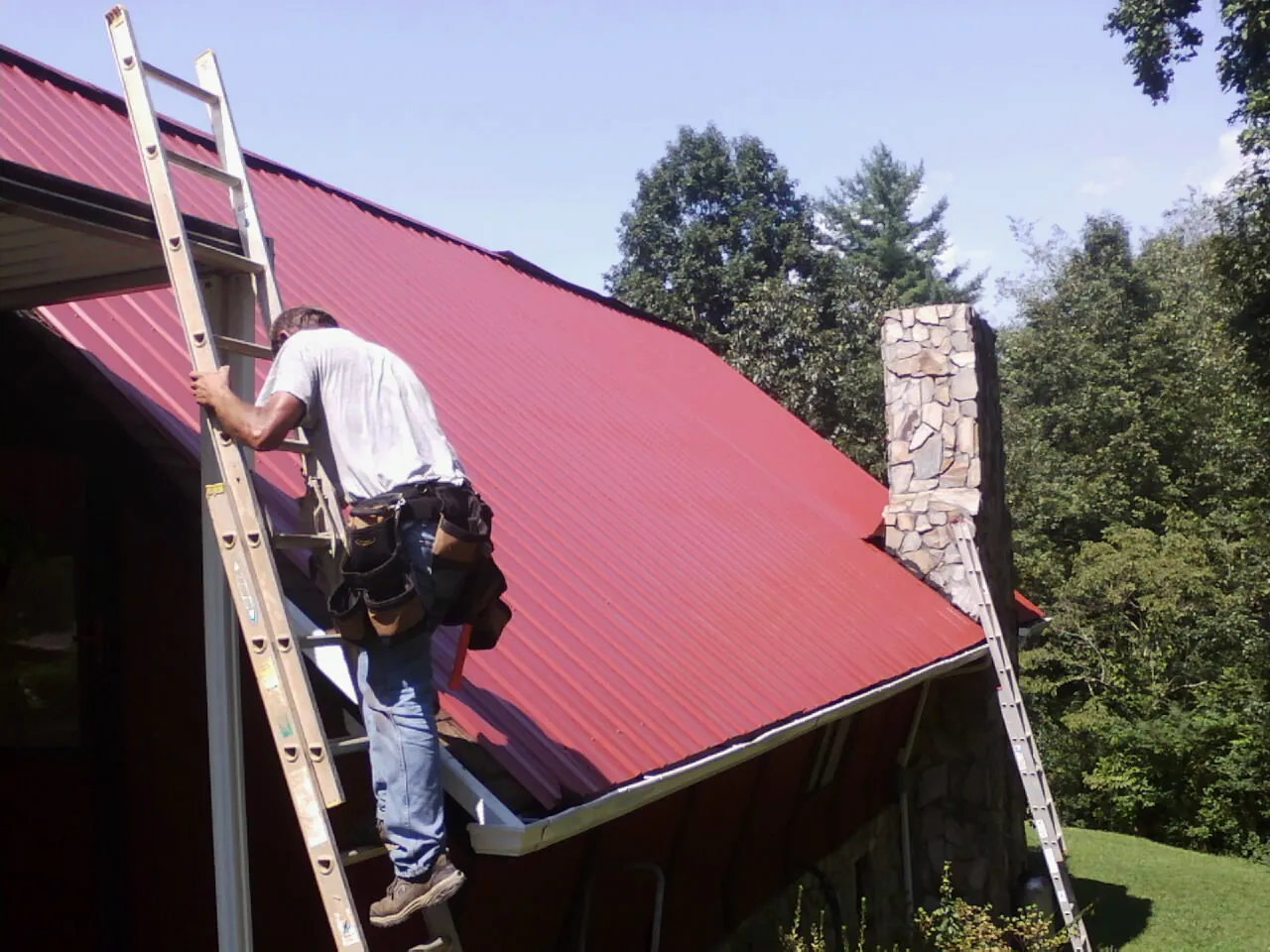 Roofer on a ladder beside a red metal roof and stone chimney — Soffit & Fascia Repair by Roofing Brothers in Hansen